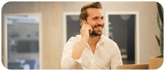 Photographie d'un homme barbu et souriant, en chemise blanche, manches remontées, qui téléphone dans un environnement de bureau.